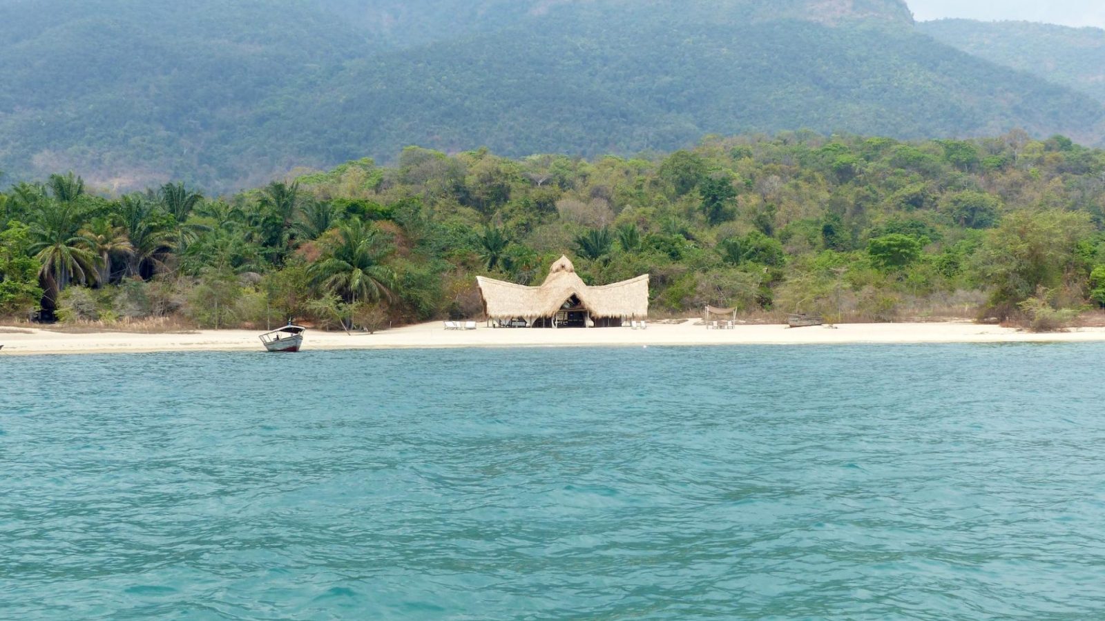 A single wooden boat on turquoise lake water below hazy forested mountains